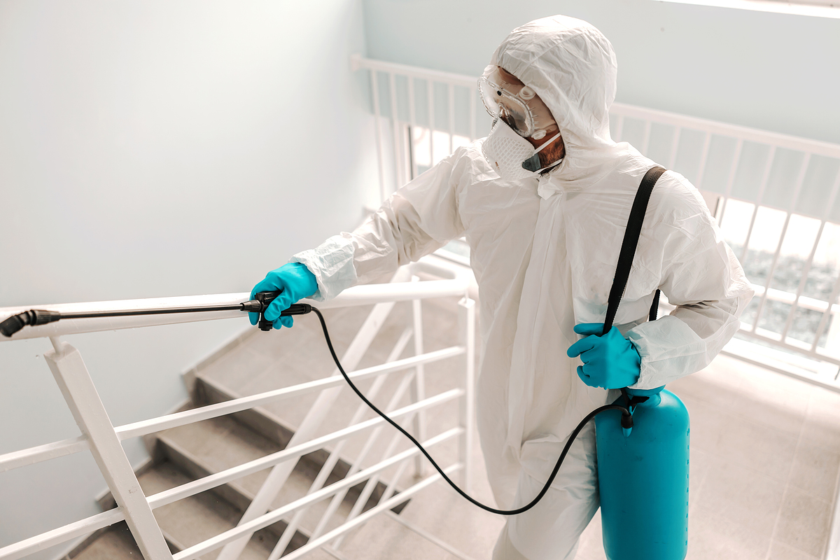 Worker in sterile uniform, with gloves and facial mask sterilizing railing in school.