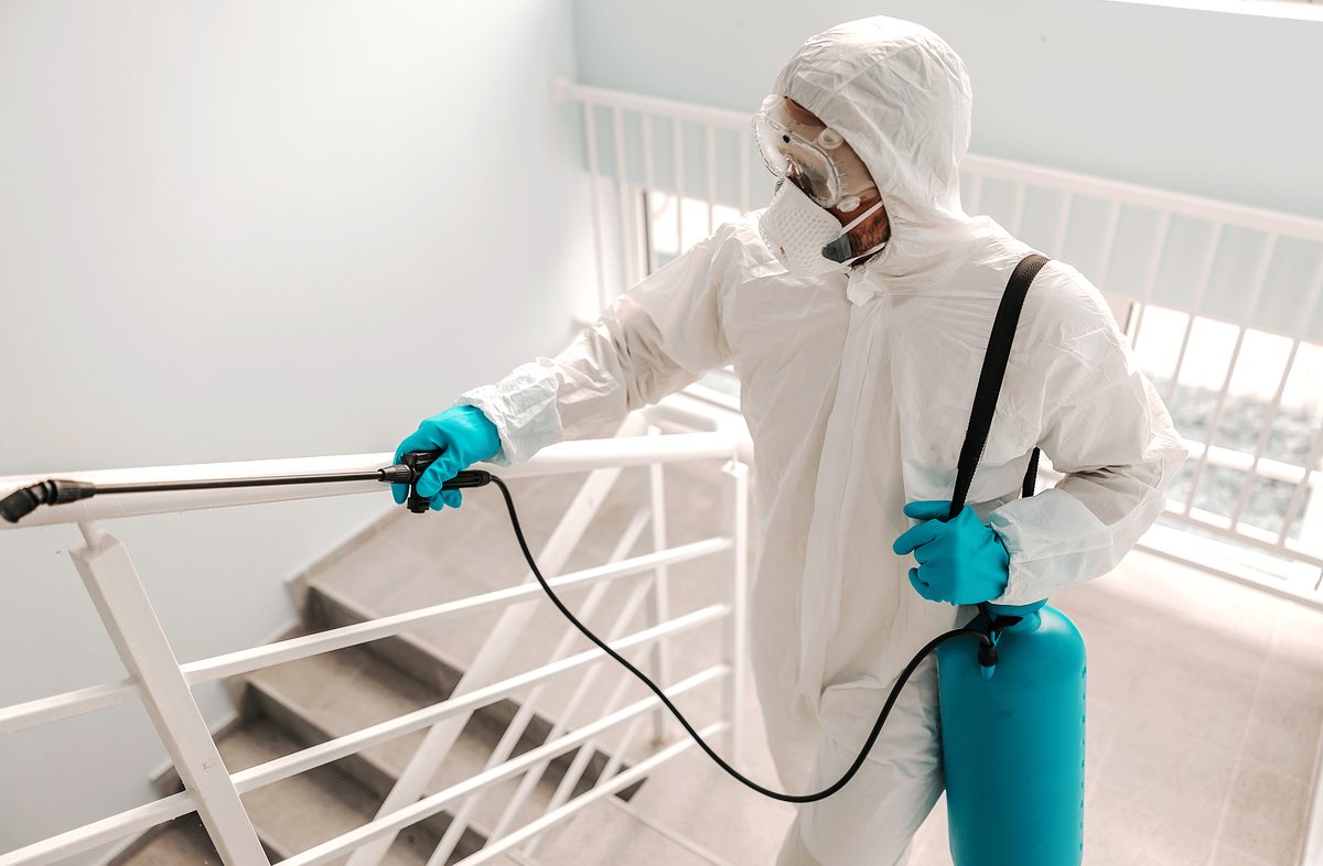 Worker in sterile uniform, with gloves and facial mask sterilizing railing in school.
